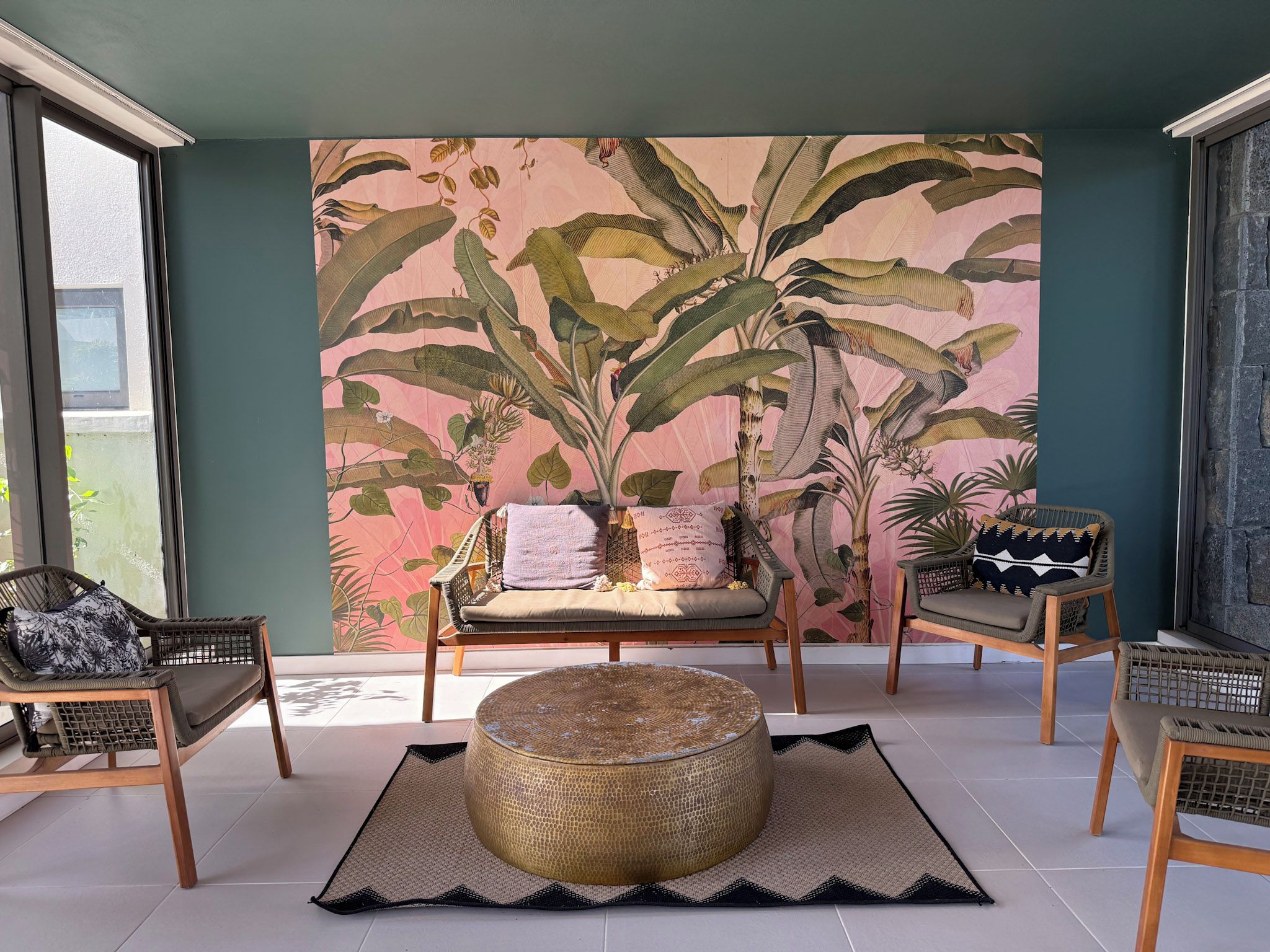 A room with a large tropical plant mural, a round wooden ottoman, and two wicker chairs.