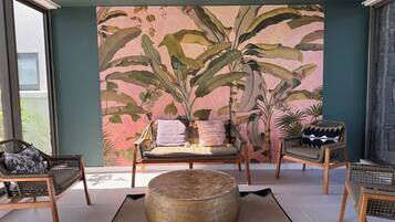 A room with a large tropical plant mural, a round wooden ottoman, and two wicker chairs.