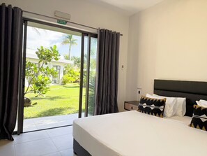 Habitación de hotel con cama, mesita de noche, almohada y vista a un jardín a través de una puerta corredera.
