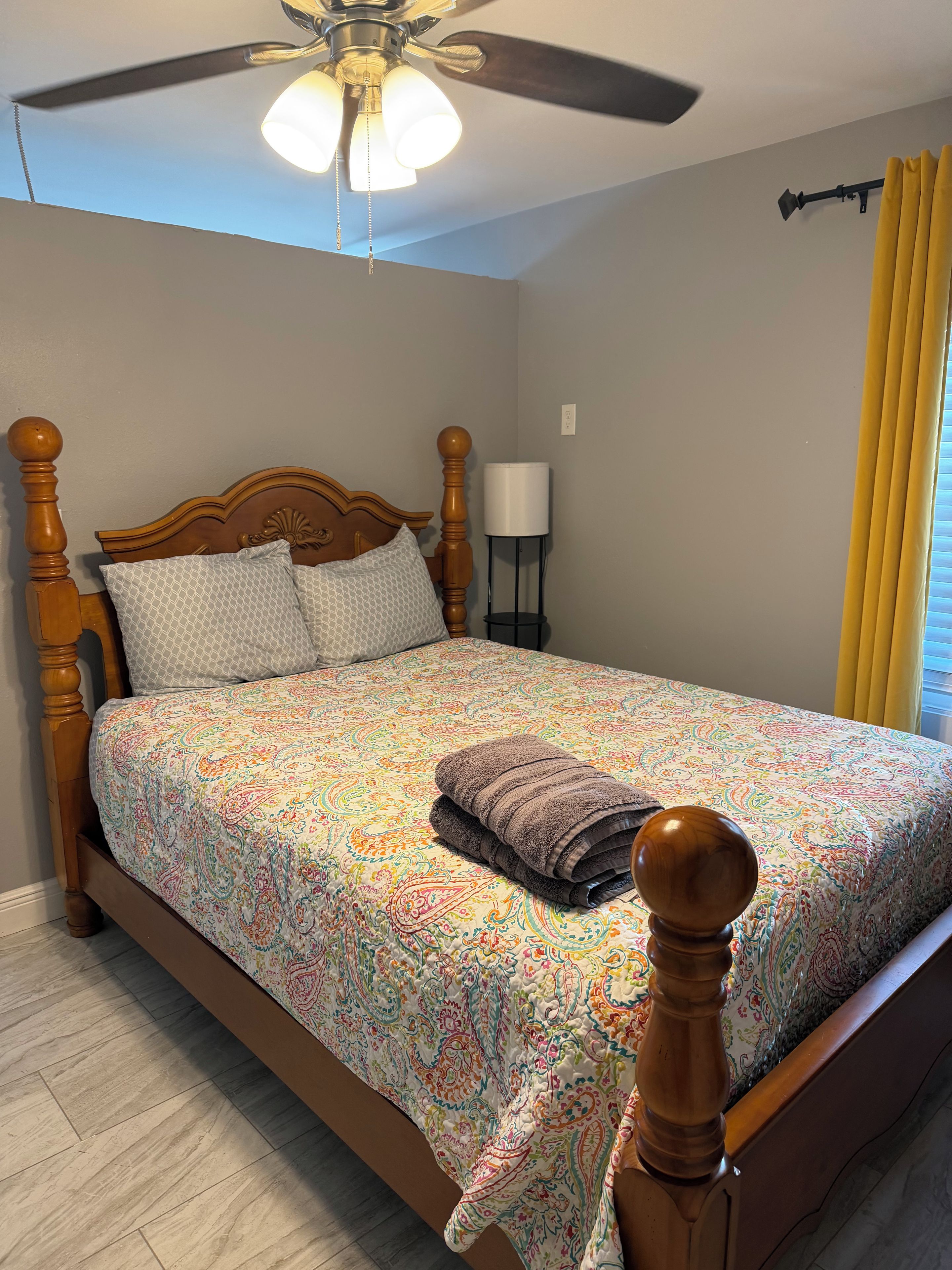 Une chambre à coucher avec un lit en bois, une literie à motifs, un ventilateur de plafond et une table de chevet avec une lampe.