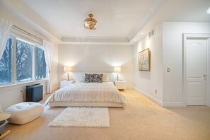 Een ruime slaapkamer met een groot bed, twee nachtkastjes, een kroonluchter, een raam met gordijnen en een deur.