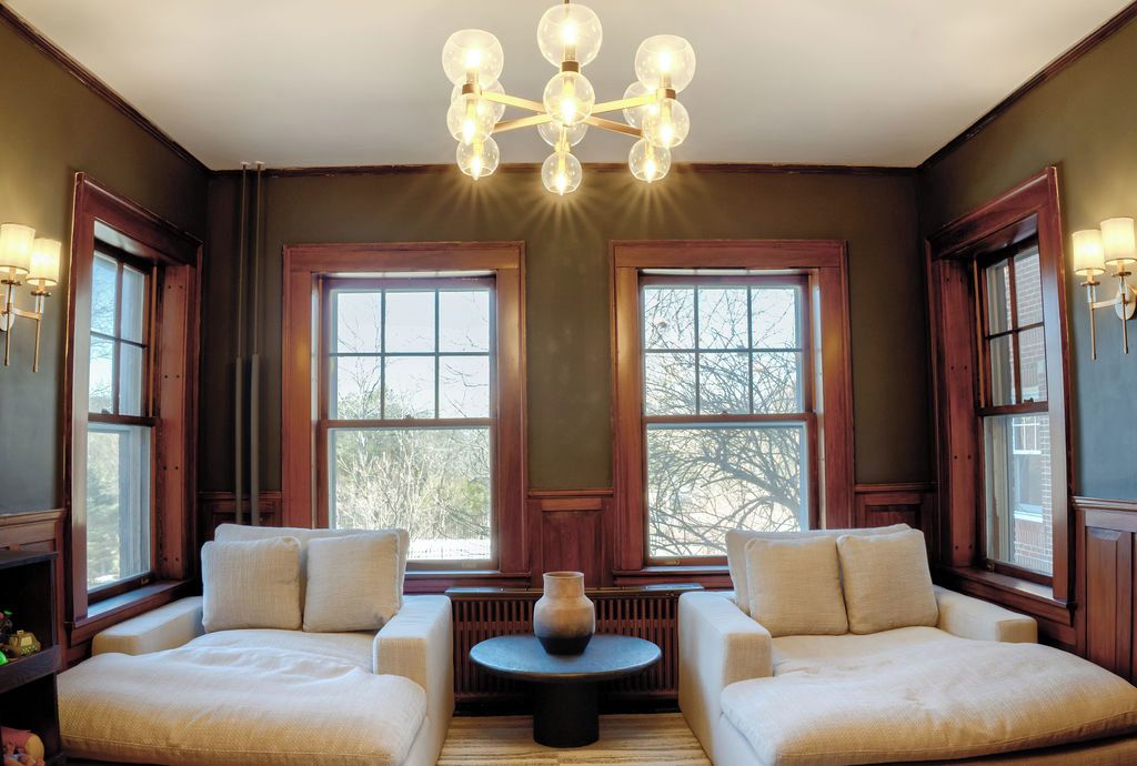 A room with two sofas facing a central table, large windows with wooden frames, and a chandelier.