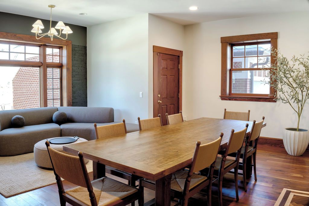A dining area with a wooden table and chairs, a sofa, a potted plant, and a window with a view of a brick building.