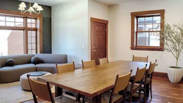A dining area with a wooden table and chairs, a sofa, a potted plant, and a window with a view of a brick building.