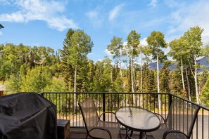 An outdoor patio with a table and chairs, surrounded by a metal railing, overlooking a forested area with tall trees and mountains in the distance. - Mountain Village Gem|Hot Tub+Views+Ski-In/Out (Mountain Village)