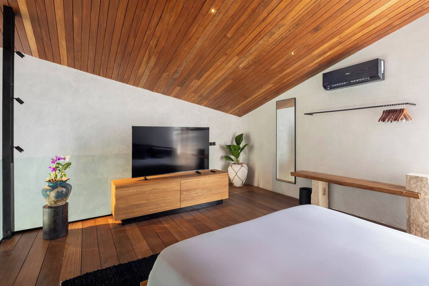 Ein modernes Schlafzimmer mit Holzboden, einem großen Flachbildfernseher, einem hölzernen Bett, einem Nachttisch, einer Topfpflanze und einer Wand-Klimaanlage.