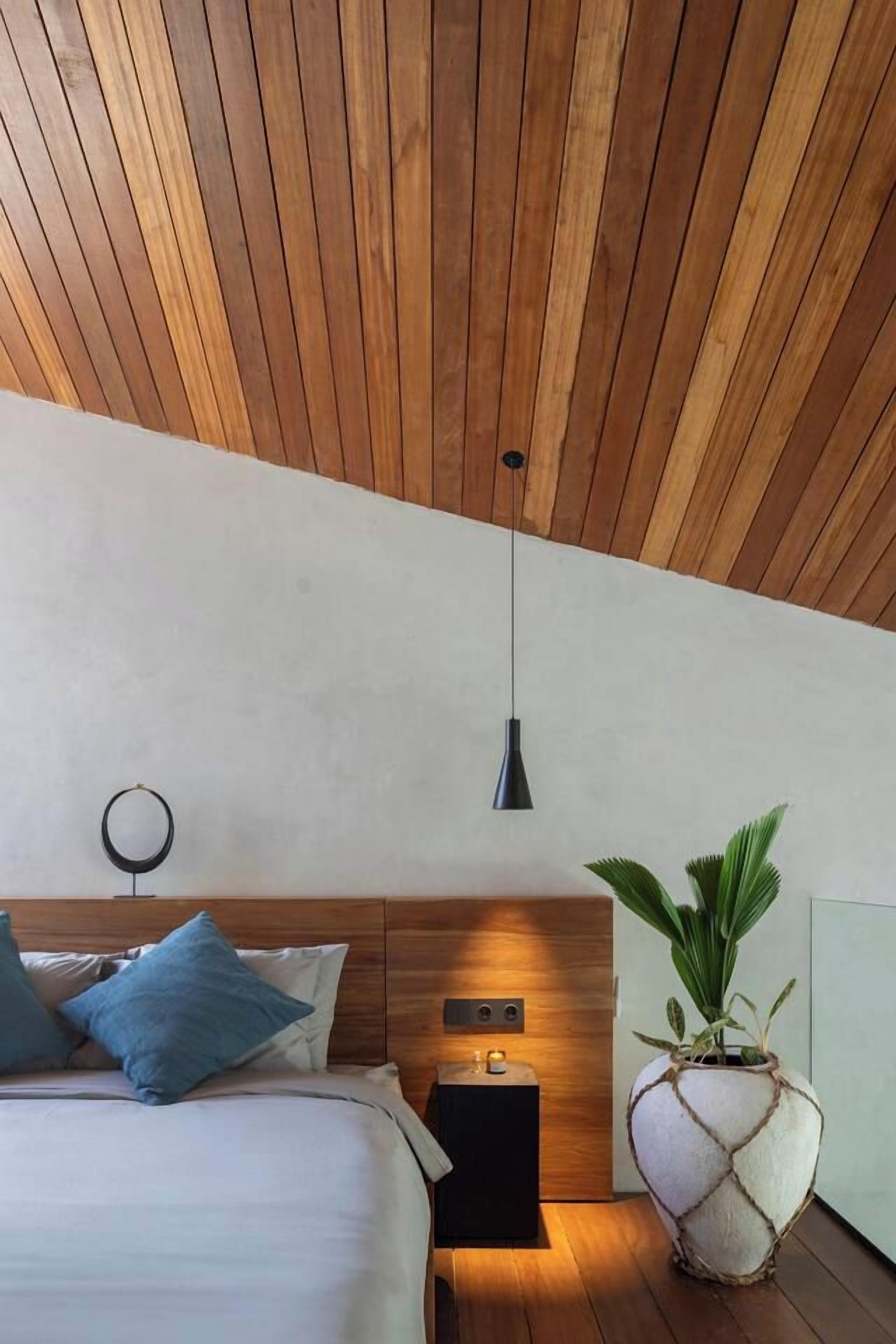 Ein modernes Schlafzimmer mit Holzbalkendecke, einem Bett mit blauen Kissen, einem Nachttisch mit Lampe und einer großen Topfpflanze.