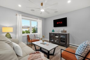 Una sala de estar con sofá, sillón, mesa de centro y televisor montado en la pared.