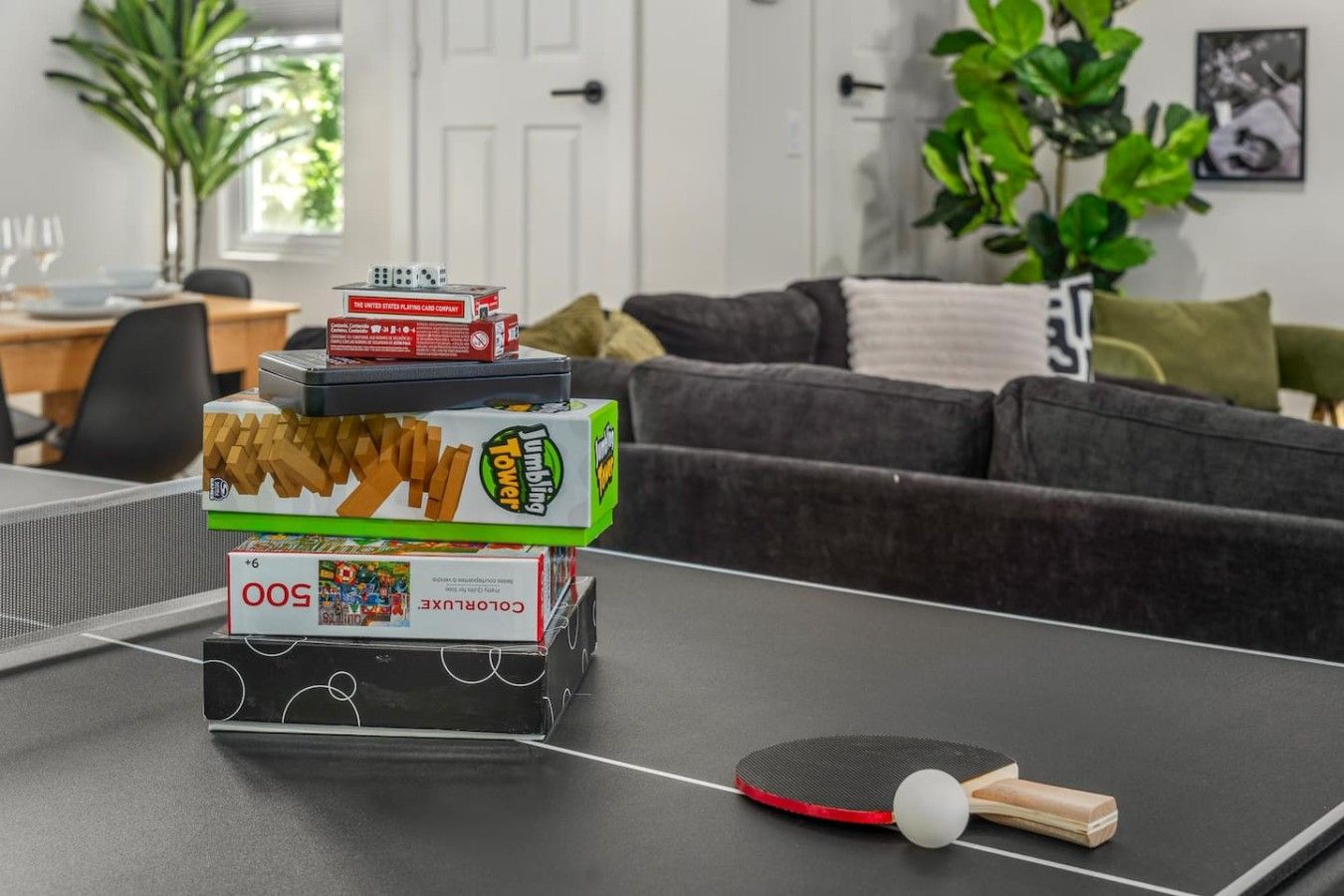 A ping pong table with a paddle and balls, a stack of board games, and a couch in a living room.