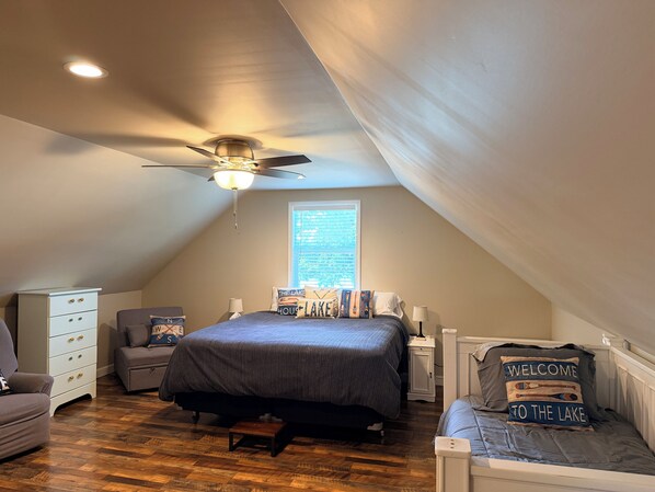 A bedroom with a sloped ceiling, a bed with a blue bedspread, a small bedside table, a chair, a nightstand, and a window with blinds.