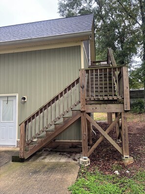 Un escalier en bois mène à une terrasse située à l’extérieur d’un bâtiment, qui est doté d’une porte et d’une fenêtre.