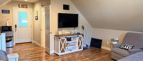 A cozy living room with a wooden floor, a ceiling fan, a flat-screen TV, and a comfortable seating area.