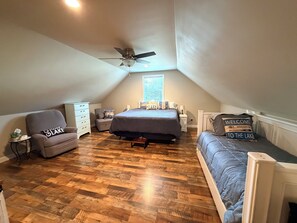 Une chambre avec un plafond en pente, un lit recouvert d’une couverture grise, une chaise, une petite table et une table de chevet.