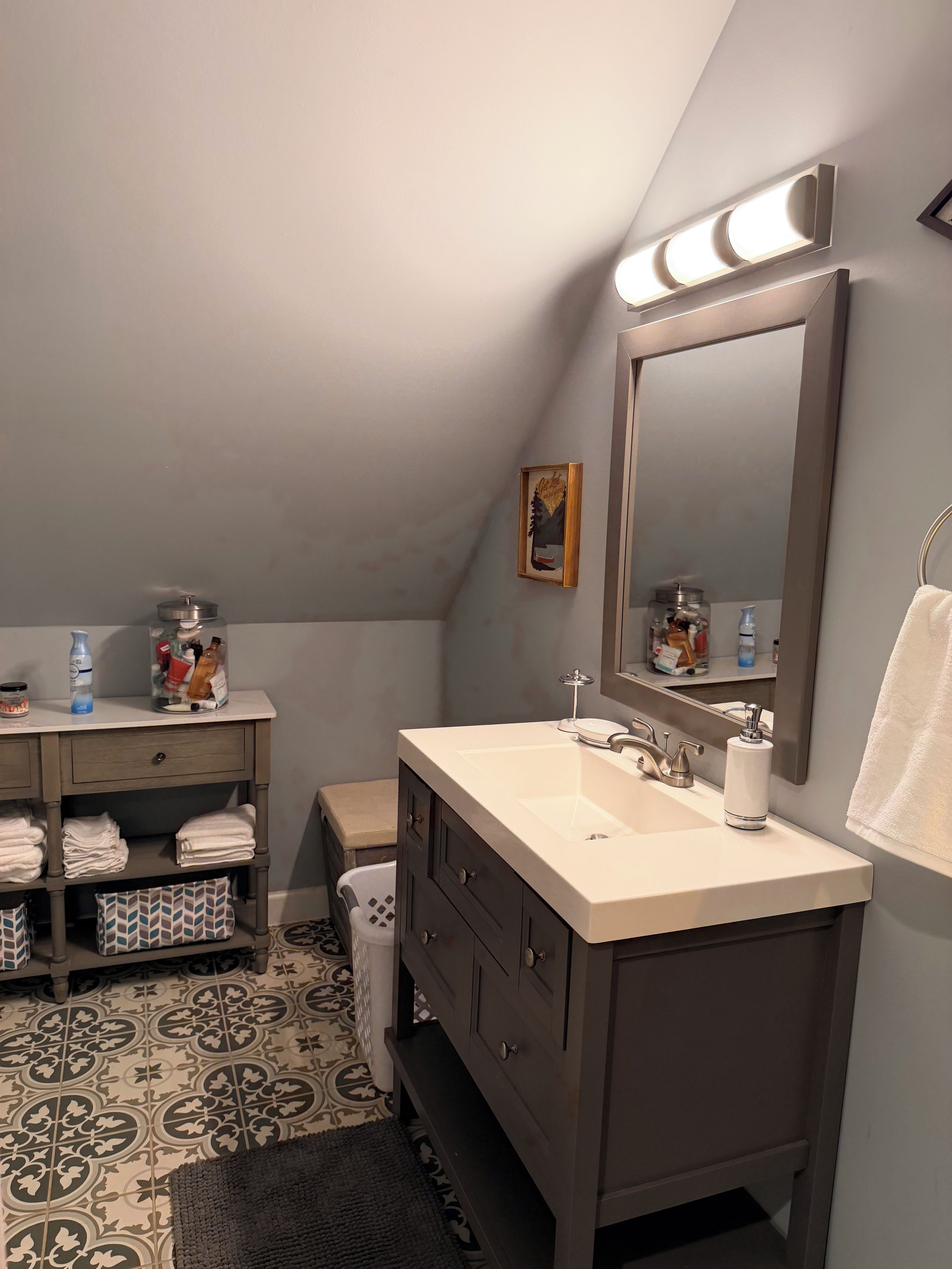 A bathroom with a vanity, sink, and mirror. There is a towel rack, a trash bin, and a shelf with toiletries.