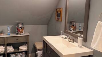 A bathroom with a vanity, sink, and mirror. There is a towel rack, a trash bin, and a shelf with toiletries.