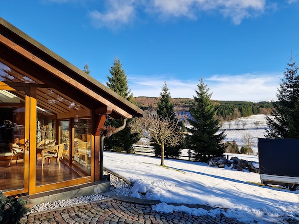 A wooden cabin with a glass entrance, outdoor seating, and a snowy landscape with trees and hills in the background.