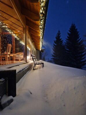 Een sneeuwbedekt terras met een bankje en houten leuning, uitkijkend op een besneeerd landschap met bomen.