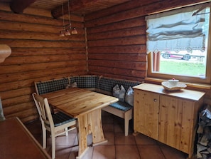 Interior de una cabaña con una mesa de madera, asientos tipo banco, un mueble pequeño y una ventana con vistas al exterior.