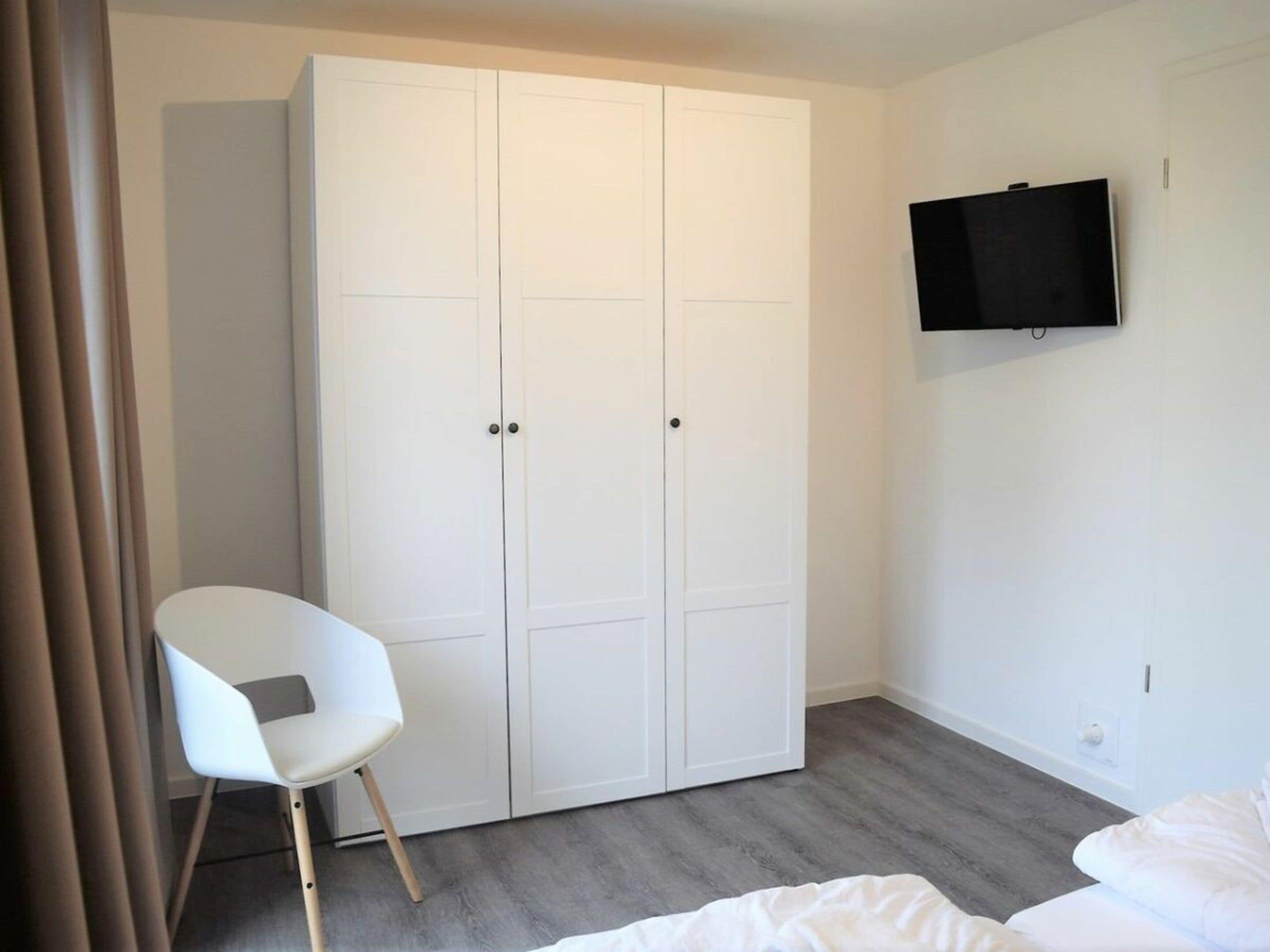 A bedroom with a bed, a white wardrobe, a flat-screen TV mounted on the wall, and a white chair.