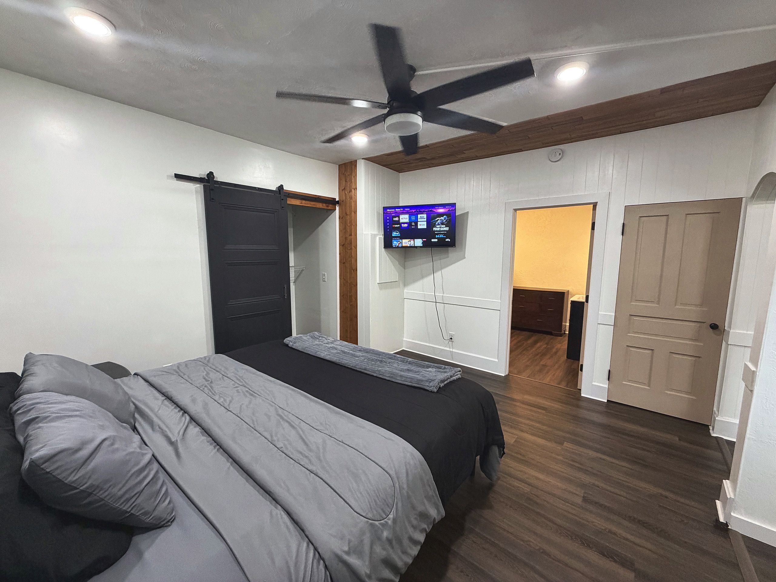 Een slaapkamer met een bed, een plafondventilator, een aan de muur bevestigde televisie en een deur naar een andere kamer.