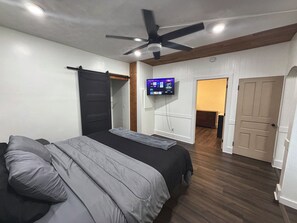 Een slaapkamer met een bed, een plafondventilator, een aan de muur bevestigde televisie en een deur naar een andere kamer.