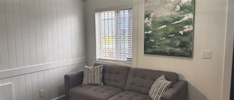 A room with a grey upholstered sofa, a small round table, a painting on the wall, and a window with blinds.