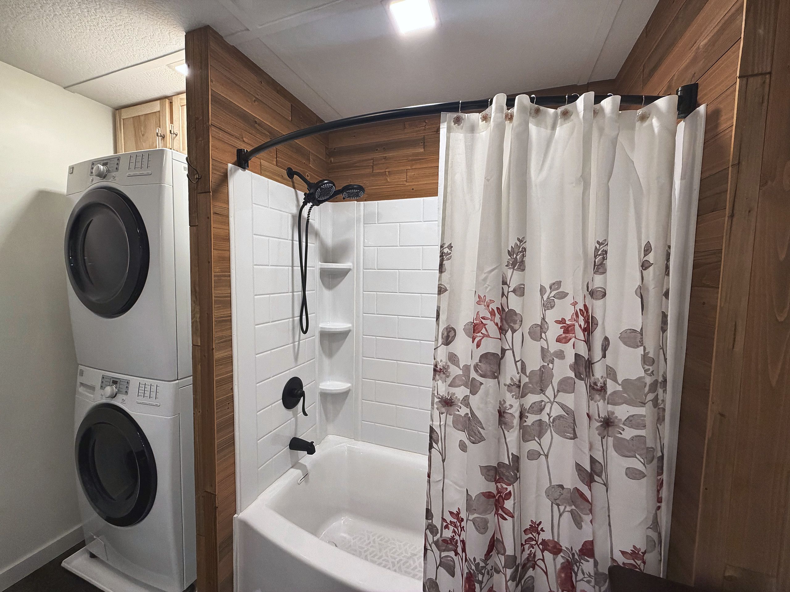 Un espace buanderie équipé d’un lave-linge et d’un sèche-linge à chargement frontal, une douche avec un mur en bois et un rideau de douche à motifs floraux.