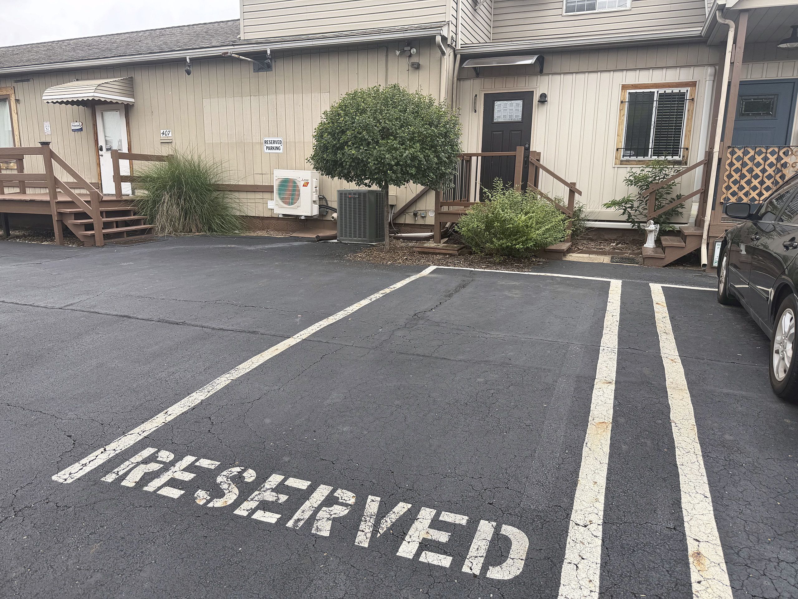 Un área de estacionamiento con la palabra 'RESERVED' pintada en el suelo, un edificio con una puerta y escalones, y un coche estacionado al lado.