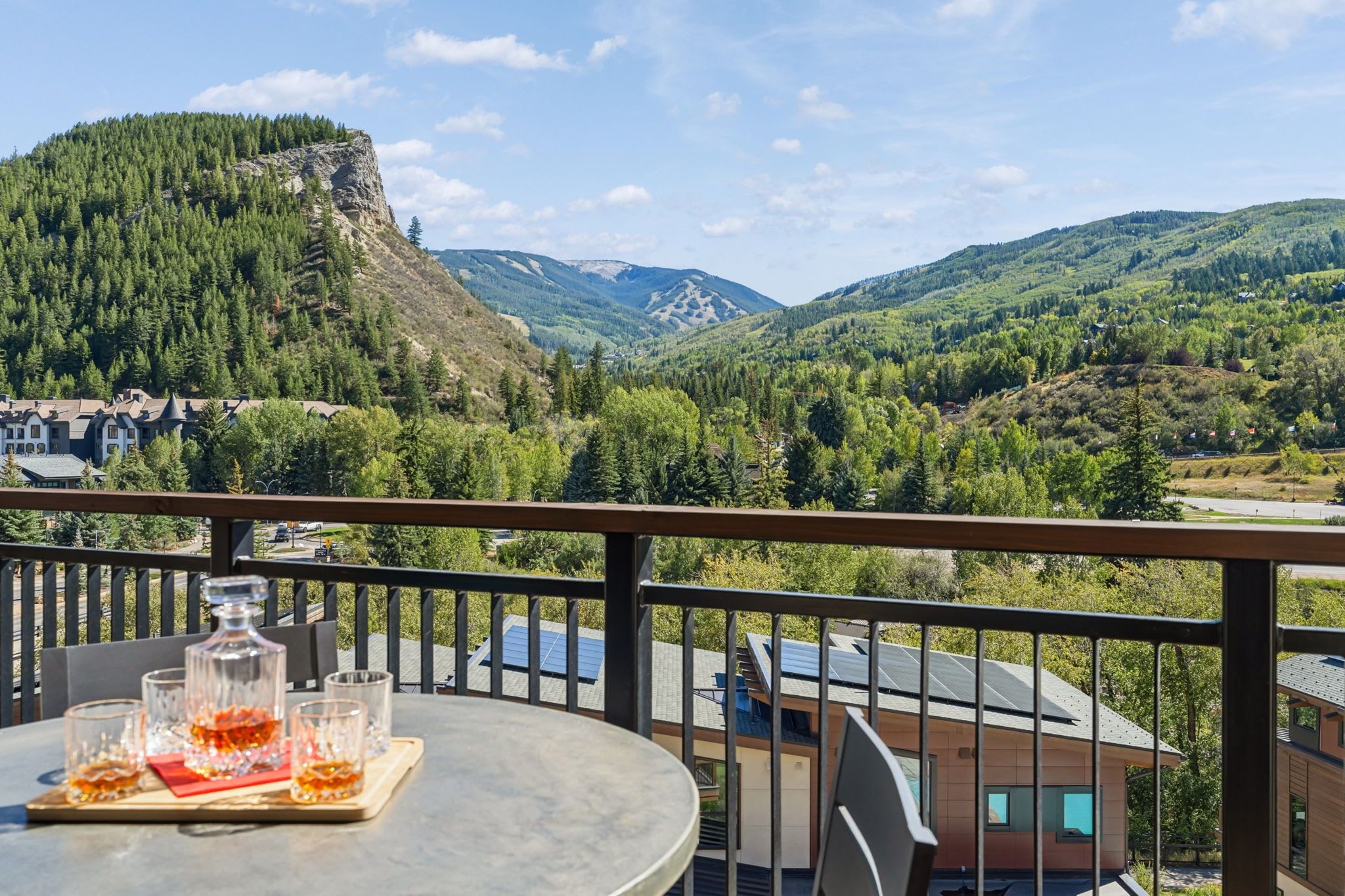 A balcony with a table set for two, overlooking a mountainous landscape with a view of a village and a prominent peak.