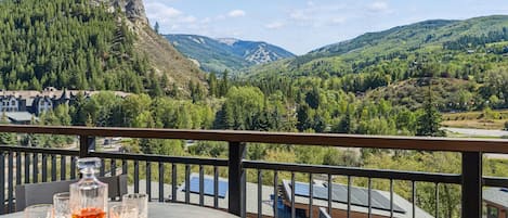 A balcony with a table set for two, overlooking a mountainous landscape with a view of a village and a prominent peak.