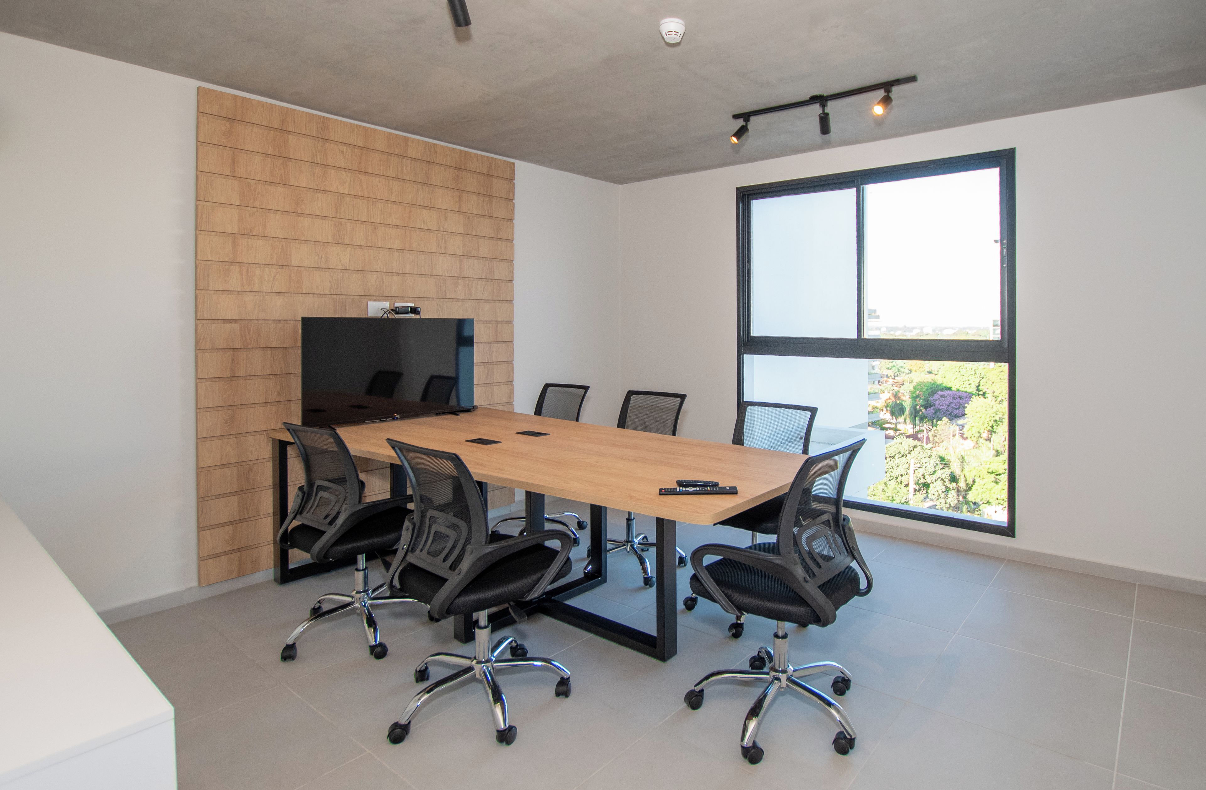 A conference room with a long table, six chairs, a television on a wooden wall, and a large window with a view of greenery.