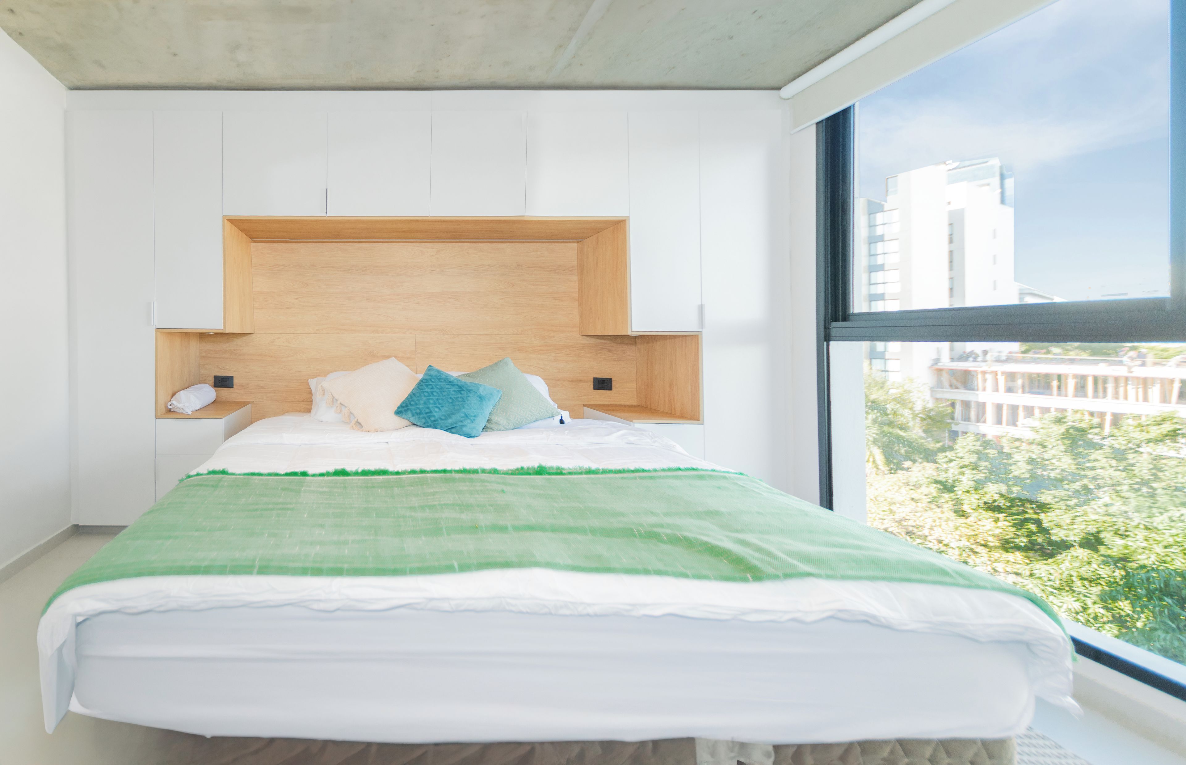 Un dormitorio moderno con cama, cabecera de madera y vistas a edificios a través de un ventanal amplio.