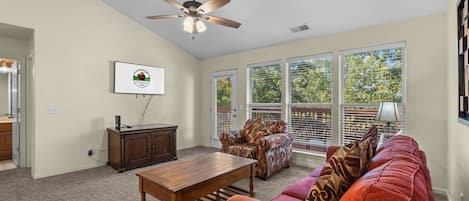 A living room with a ceiling fan, a television mounted on the wall, a wooden cabinet, a red sofa, and a patterned armchair.