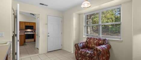 Una habitación con un sillón estampado, una ventana con persianas y una cocina visible a través de una puerta abierta.