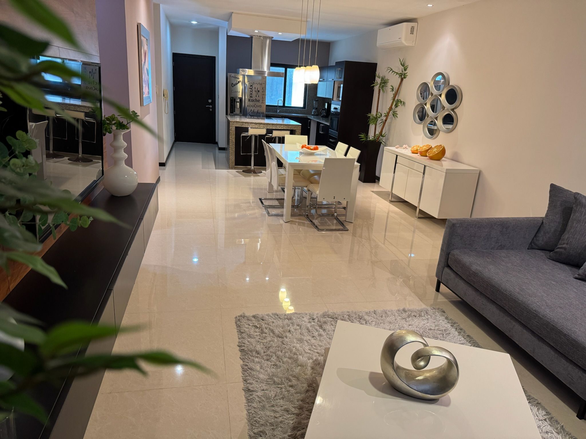 Een moderne woonkamer met een grijze bank, een eethoek met een glazen tafel en een keuken met een zwart aanrecht.