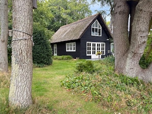 Ein schwarzes Haus mit Strohdach, umgeben von Bäumen und einem grünen Rasen.