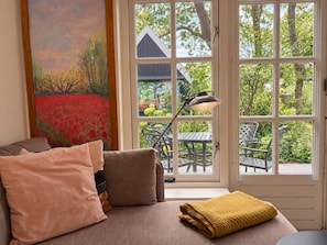 A room with a sofa, a painting, a window with a view of a garden, and a table with chairs outside. - Unique Artist’s Atelier in Bergen | Near Dunes & Beach (Bergen)