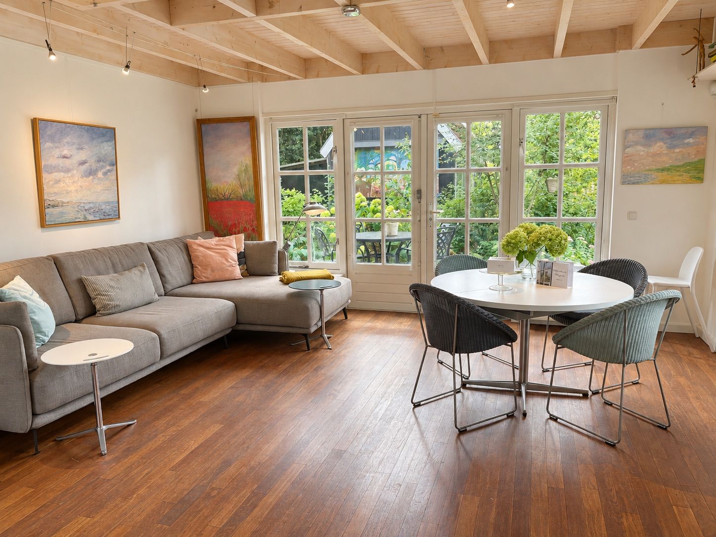 A modern living room with a grey sectional sofa, a round dining table with chairs, and large windows offering a view of greenery.
