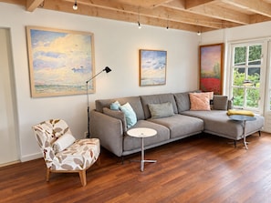 Ein modernes Wohnzimmer mit einem grauen Ecksofa, einem gemusterten Sessel und einem kleinen runden Tisch. An den Wänden hängen drei Bilder und ein großes Fenster bietet Blick auf Grünflächen.
