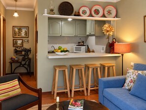 Ein Wohnzimmer mit blauem Sofa, Holzschemeln, einem Couchtisch mit einem Buch und einer Küchenzeile mit Mikrowelle und Obstschale.