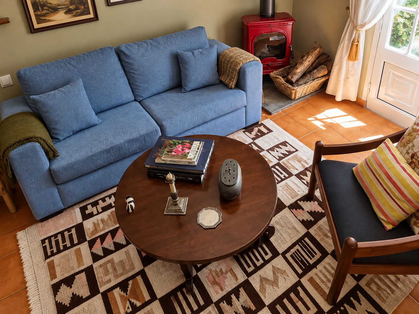 A living room with a blue sofa, a round wooden coffee table with decorative items, a patterned rug, and a red wood-burning stove.