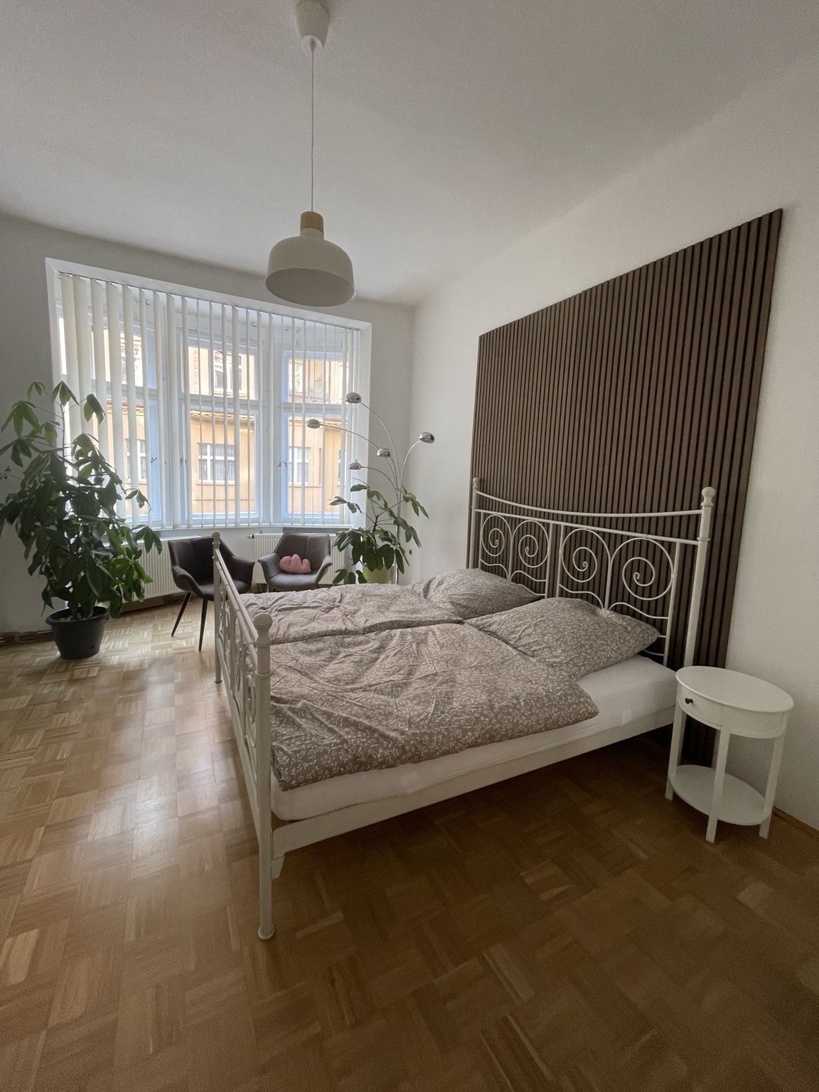 Ein Schlafzimmer mit einem Bett, einem Nachttisch, einer Zimmerpflanze, einem Sessel und einem Fenster mit Jalousien.