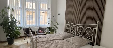 A bedroom with a bed, a bedside table, a potted plant, a chair, and a window with blinds.