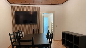 Uma área de refeições com uma mesa comprida, cadeiras, uma TV de tela plana fixada em uma parede e um armário embutido.