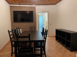 Een eetgedeelte met een lange tafel, stoelen, een flatscreen televisie aan een wand en een inbouw wandplank.