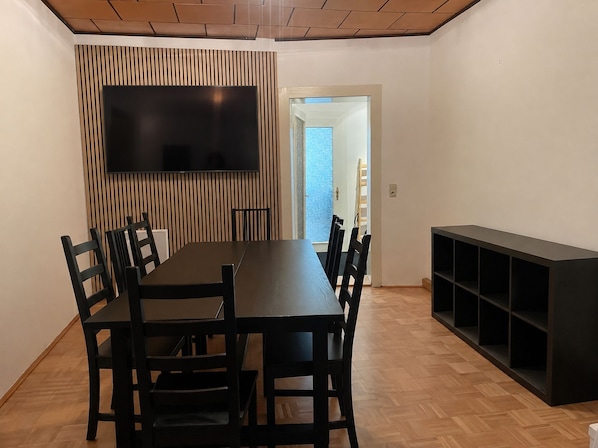 A dining area with a long table, chairs, a flat-screen TV mounted on a wall, and a built-in shelving unit.