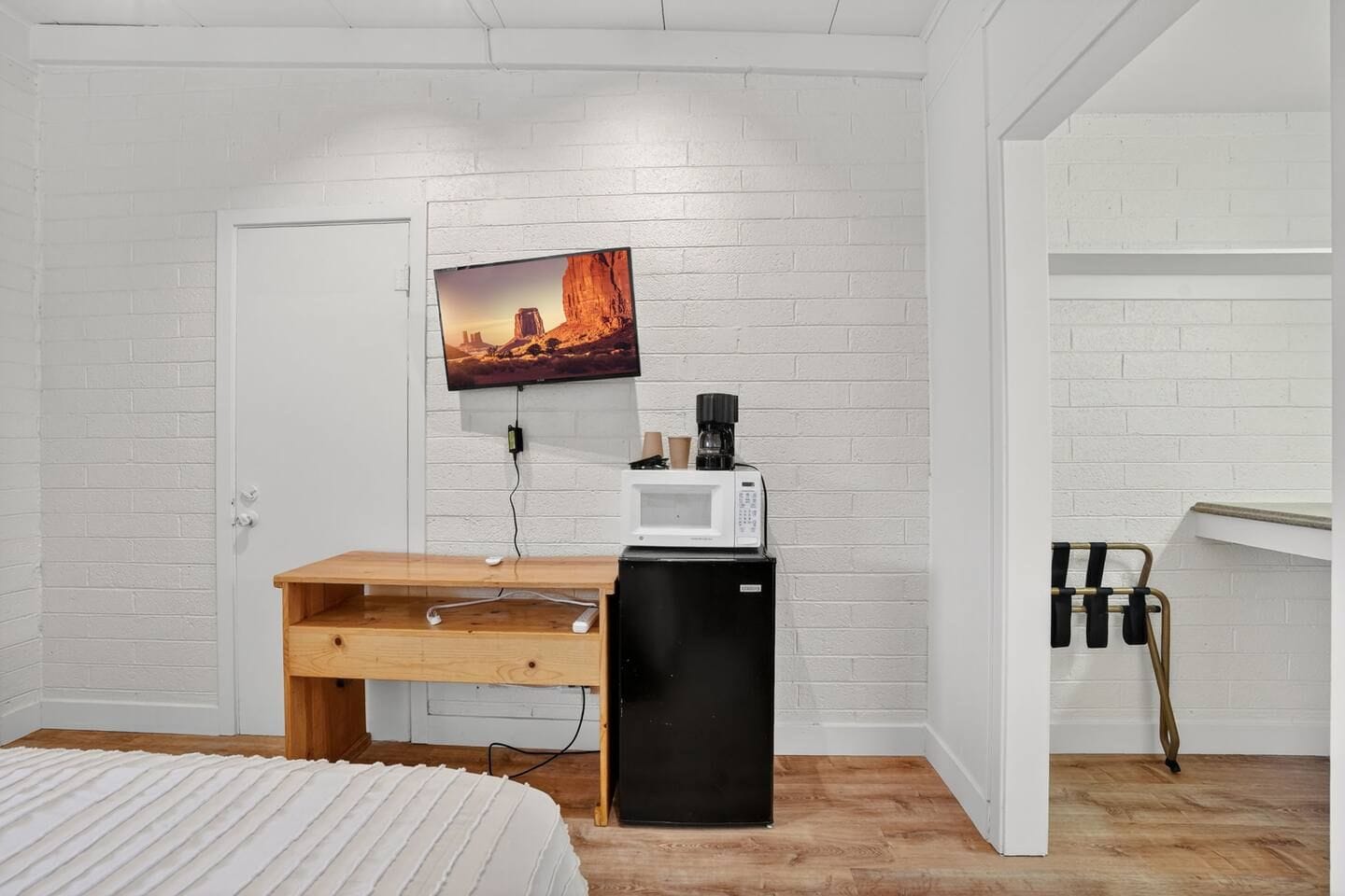 A bedroom with a white brick wall, a bed with a white bedspread, a wooden desk, a black refrigerator, a television mounted above the desk, and a white door.