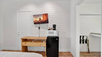 A bedroom with a white brick wall, a bed with a white bedspread, a wooden desk, a black refrigerator, a television mounted above the desk, and a white door.