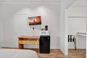 Un dormitorio con pared de ladrillo blanco, cama con colcha blanca, escritorio de madera, refrigerador negro, televisor montado sobre el escritorio y una puerta blanca.