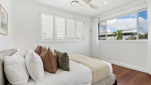 A bedroom with a bed, white shutters, a ceiling fan, and a large window with a view of buildings and sky. - The North Point Mansion (Kingscliff)